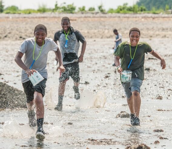 The image shows three young boys running through shallow water. They are all smiling and appear to be enjoying themselves. They are wearing casual clothes and lanyards around their necks. In the background, there is another person. The scene seems to be outdoors on a sunny day.